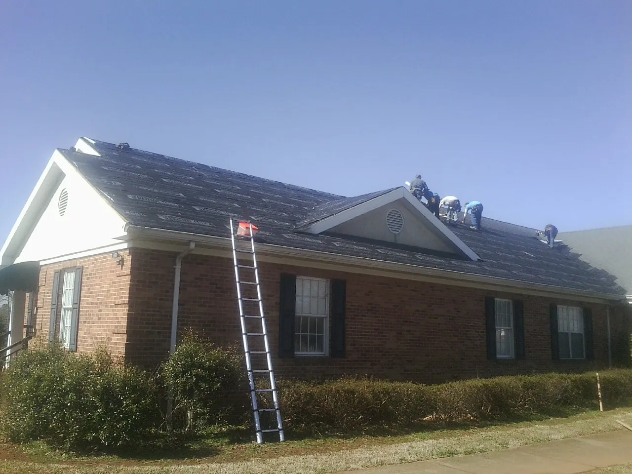 Crew tearing shingles off a brick ranch roof with a ladder set — Emergency Roof Repair work by our crew in Lidgerwood
