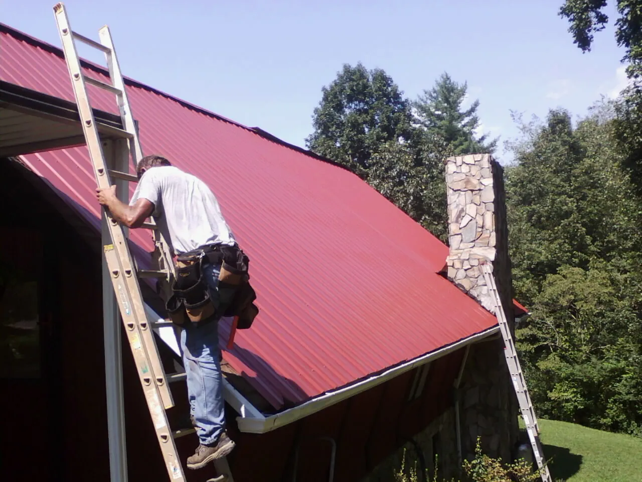 Roofer on a ladder beside a red metal roof and stone chimney — Soffit & Fascia Repair by Roofing Brothers in Lidgerwood